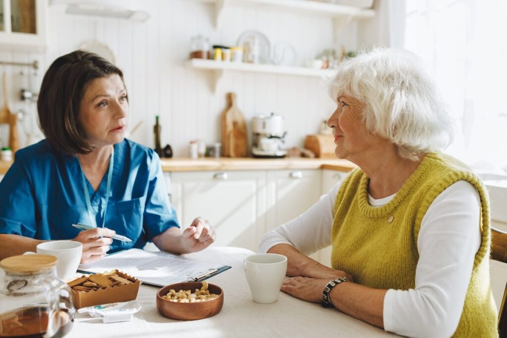 Elderly woman with grey hair answering questions of female social worker sitting at kitchen table drinking tea as first acquaintance before providing caregiver support program for aged people